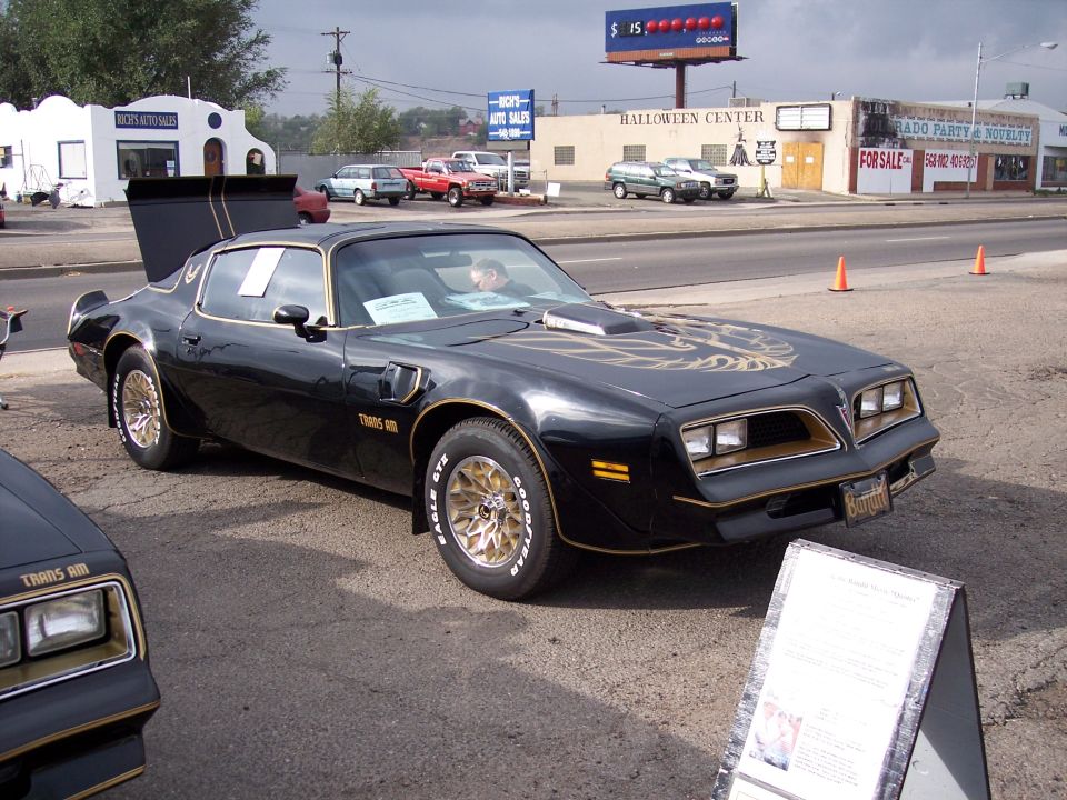 Black 1977 Pontiac Trans Am with gold decals and gold wheels, another Trans Am front end visible on left