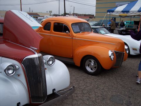 1972 Ford Ranchero - An orange hot rod coupe is parked at an outdoor car show next to other classic vehicles.