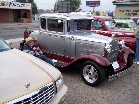1972 Ford Ranchero - A woman knits while sitting in a vintage silver and maroon coupe, parked alongside other vehicles at a show.