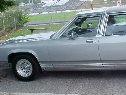 1987 Ford Crown Victoria LTD - A silver 1980s Ford sedan is parked outdoors, with stadium bleachers in the background.