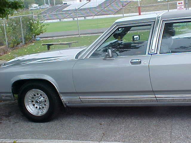 Silver box-body full-size sedan in side profile with aluminum mesh wheels, bleachers behind