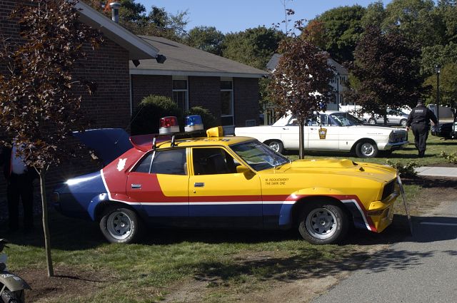 Yellow and blue police car with red stripe, light bar, and custom hood scoop. White sedan in background