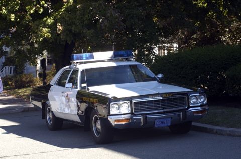 1973 Dodge 1500 - A black and white police car, marked "Deputy Sheriff", is parked on a street with trees and bushes in the background.