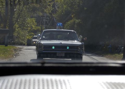 1973 Dodge 1500 - A white police car with emergency lights on drives down a tree-lined road, with another vehicle visible behind it.