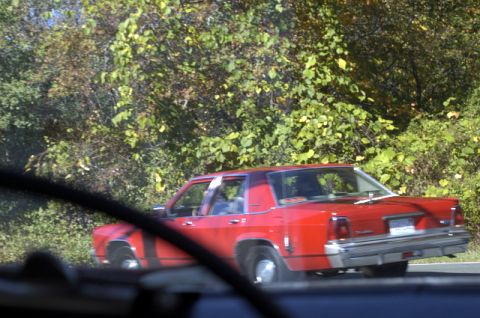 1973 Dodge 1500 - A red sedan is seen from inside another vehicle, driving on a road surrounded by trees.