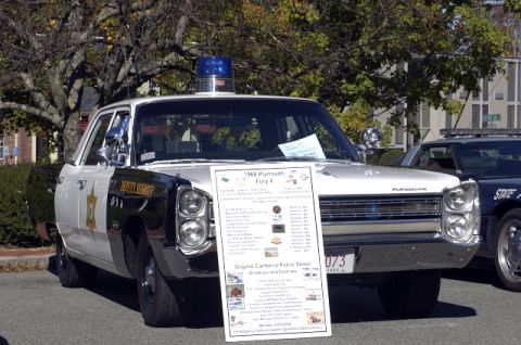 1973 Dodge 1500 - A 1968 Plymouth Fury II police car is displayed with a sign detailing its features, with another police vehicle in the background.