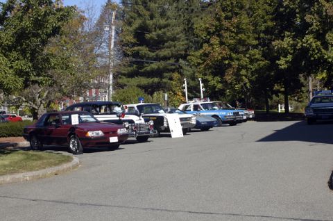 1973 Dodge 1500 - A line of police cars and a maroon coupe are parked on asphalt under trees.