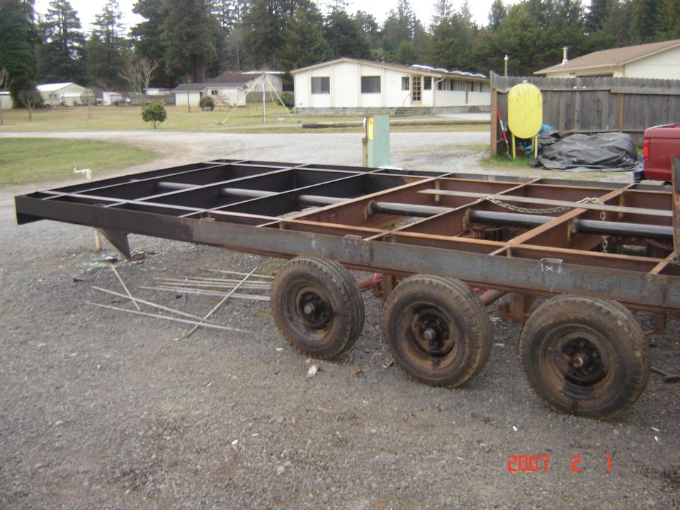 Unfinished trailer frame with multiple axles and wheels, parked on gravel