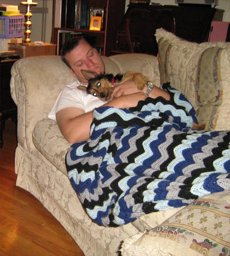 Man napping on a couch holding a small dog under a blanket