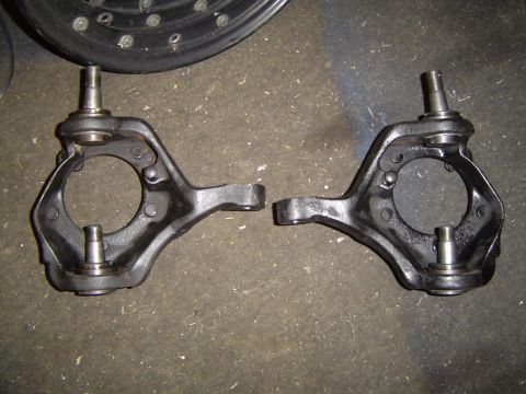 1978 Ford Bronco - Two black steering knuckles, components of a vehicle's suspension system, are displayed on a rough surface.