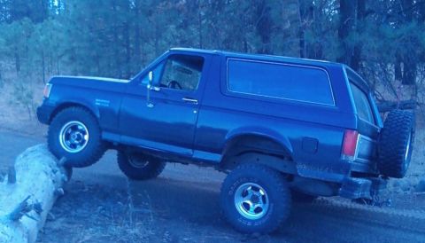1988 Ford Bronco - Passenger-side profile of a lifted blue full-size Ford Bronco with hardtop, its front tire climbing a fallen log at the edge of a dirt road…