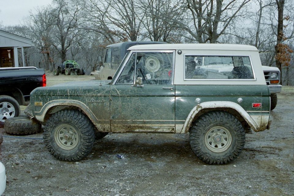 Muddy dark green Ford Bronco with white top and flares, side view on gravel lot