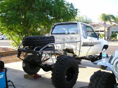 1986 Toyota Hilux - Rear three-quarter view of a bed-delete white Toyota pickup in a driveway, its bed replaced by a bare-steel tube rear section with a flat-t…