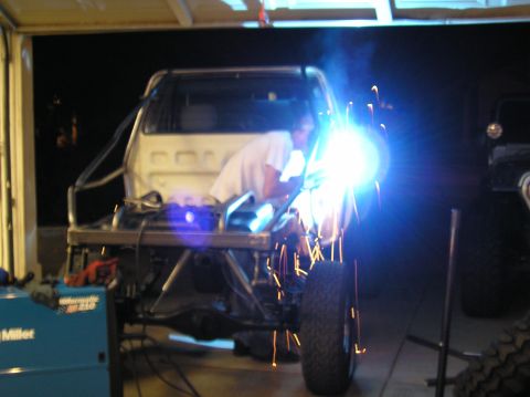 1986 Toyota Hilux - Night shot of a builder MIG-welding tube on the bare rear frame of a stripped pickup in a garage, sparks and arc glare lighting the shop. T…