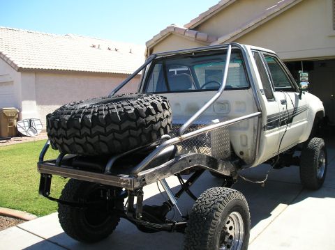 1986 Toyota Hilux - Three-quarter rear view of a white lifted mini-truck buggy build parked in a residential driveway, with the bed replaced by a bare-steel tu…