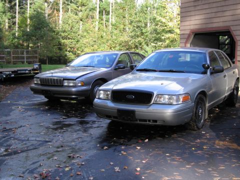 1991 Ford LTD Crown Victoria - A silver Ford Crown Victoria and a gray sedan are parked on a wet driveway next to a building.