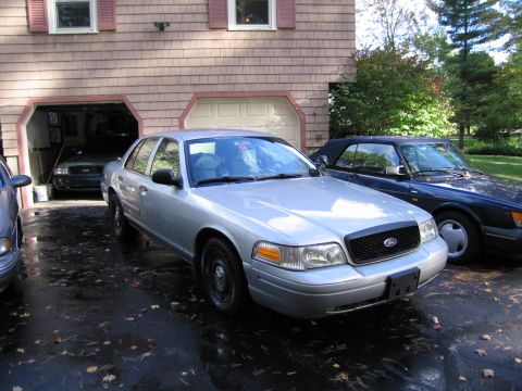 1991 Ford LTD Crown Victoria - A silver Ford Crown Victoria is parked in front of a garage, with a dark blue convertible to its right.