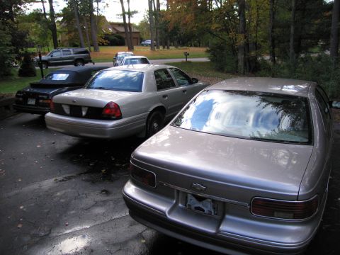 1991 Ford LTD Crown Victoria - Several vehicles are parked on a wet driveway in front of a wooded area with houses in the distance.