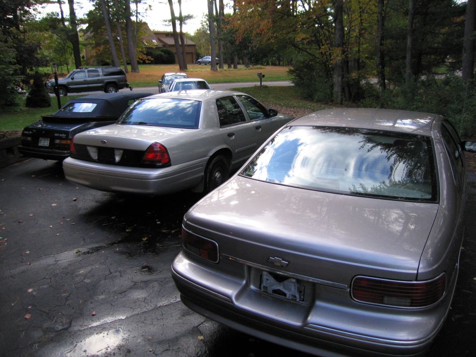 Rear view of a silver sedan parked on a wet driveway next to a black convertible and another silver sedan. An SUV is visible in the background