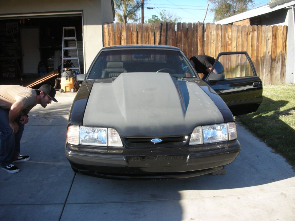 Front view of a black Fox-body Mustang with primer hood in a driveway