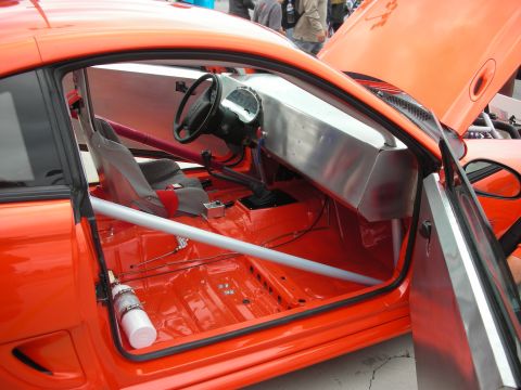 1998 Ford Crown Victoria - The interior of an orange race car is visible with its door open, showing a roll cage and racing seat.
