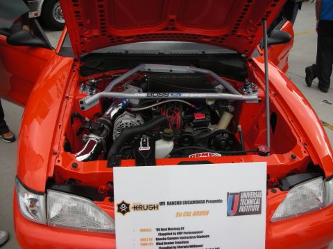 1998 Ford Crown Victoria - The engine bay of an orange Ford Mustang GT is displayed, with a sign detailing its specifications.