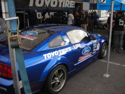 1998 Ford Crown Victoria - A blue Ford Mustang race car with sponsor decals is parked at an event.