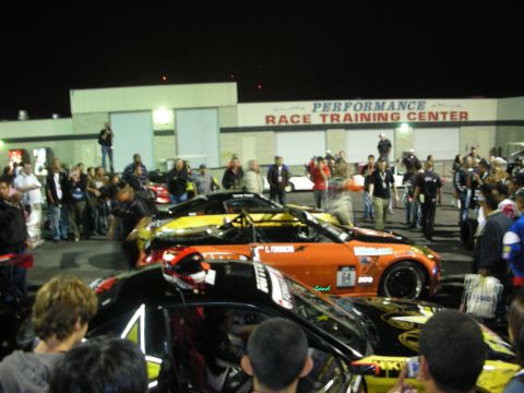 1998 Ford Crown Victoria - A crowd gathers around several colorful race cars parked in front of a building labeled 'Performance Race Training Center'.