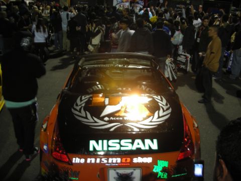 1998 Ford Crown Victoria - Orange Nissan 350Z with NISMO shop branding and racing decals at an event.