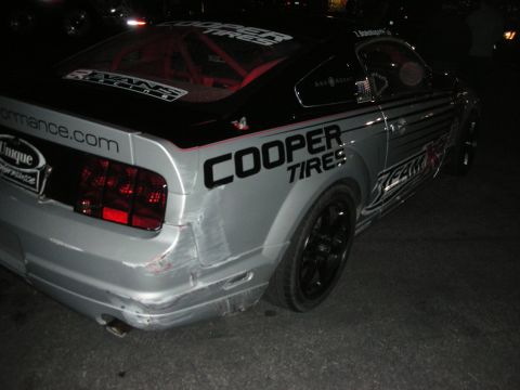 1998 Ford Crown Victoria - This silver Ford Mustang, likely a 2005 model, displays 'Cooper Tires' branding and a racing roll cage, with visible damage to the rear bum…