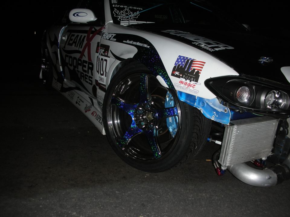 Front corner of a white sponsor-liveried sports car at night with a glitter-finish black wheel