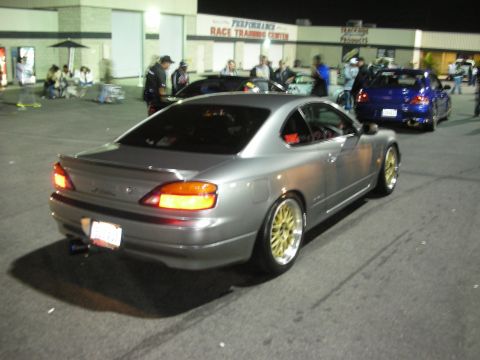 1998 Ford Crown Victoria - A silver coupe with gold rims is parked in a lot near a building with a sign that reads 'Performance Race Training Center'.