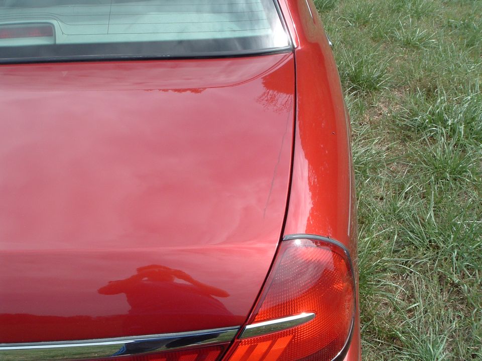 Red car's rear quarter panel, trunk lid, and taillight. Grass in background