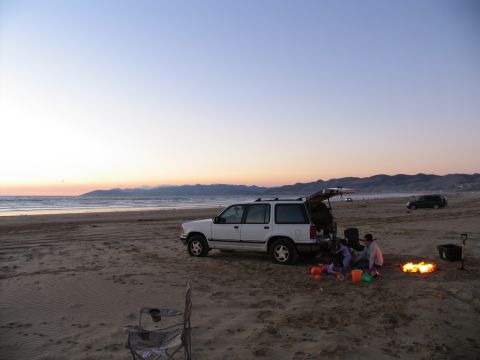 1984 Ford Bronco - Family camping on the beach with a white SUV and a campfire at sunset.