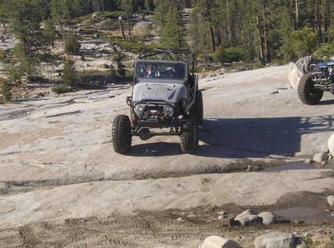 1986 Ford Bronco - Two off-road vehicles navigate a rocky, wooded landscape.