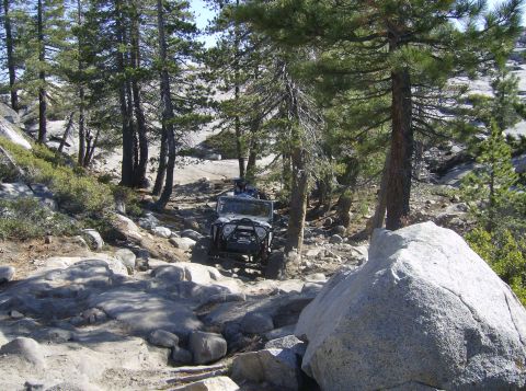 1986 Ford Bronco - A white Jeep CJ-7 tackles a challenging off-road path through a forest with large boulders.