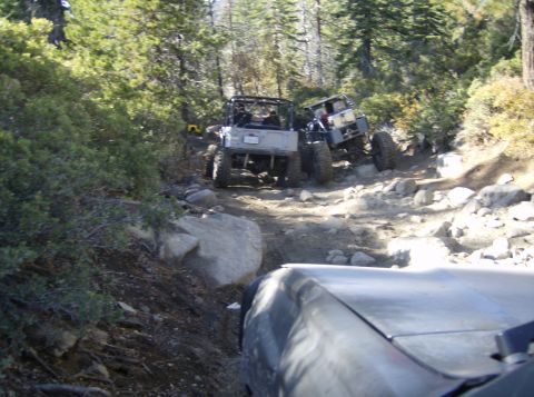 1986 Ford Bronco - Multiple off-road vehicles tackle a challenging rocky trail in a forest setting.