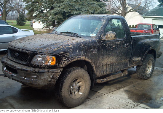 Black Ford F-150 covered in mud, parked on a concrete driveway. The truck is a regular cab with a short bed