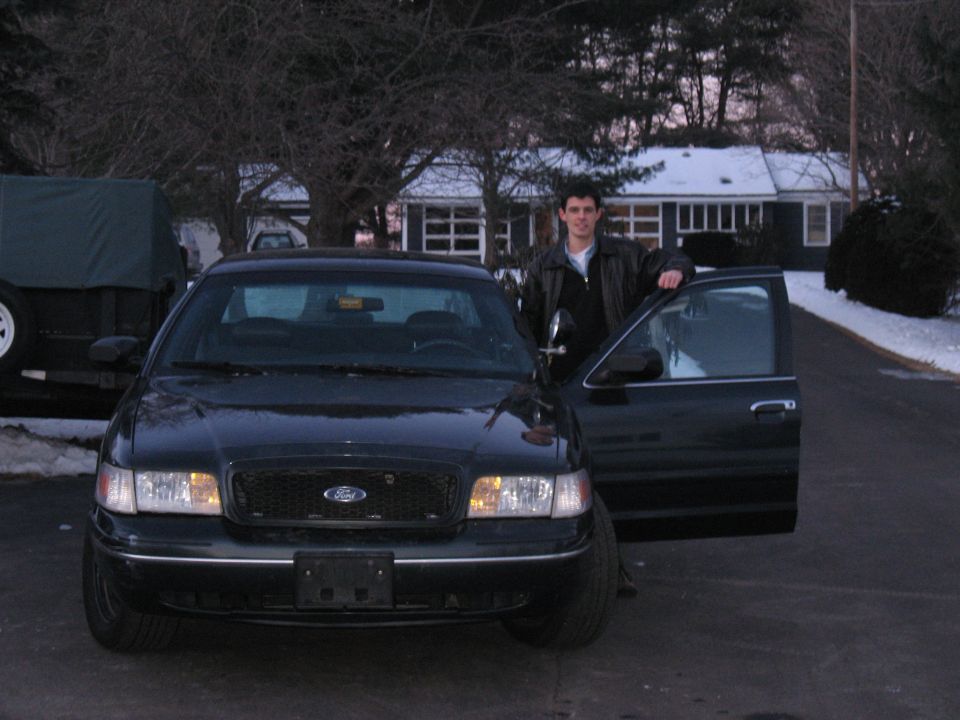 Dark green Ford Crown Victoria sedan from the front, owner standing at open driver door, snowy yard behind