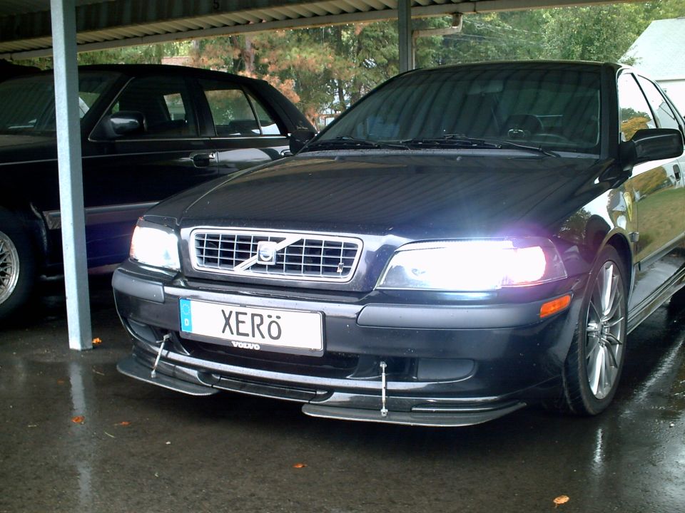 Dark Volvo S40 sedan, front 3/4 view, headlights on, 'XERö' plate, wet pavement