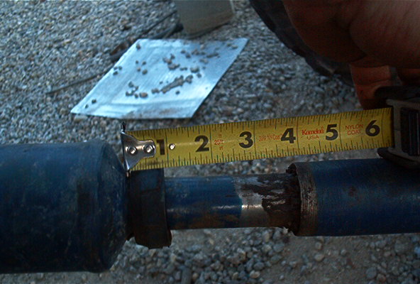 Driveshaft part being measured with a yellow tape measure against a gravel background