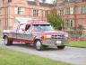 1989 Ford Bronco - A red and white pickup truck with a camper shell is parked on a paved surface with a building in the background.