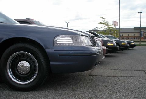 1998 Ford Crown Victoria - Low-angle view down a row of Ford Panther-body sedans parked nose-out in a shopping-center lot, with a Staples store sign in the background…