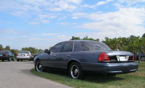 1998 Ford Crown Victoria - A dark blue-gray Ford Crown Victoria full-size sedan viewed from the rear three-quarter, parked on grass beside a paved lot with chrome-hub…
