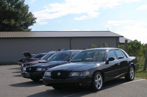 1998 Ford Crown Victoria - A row of black full-size Mercury sedans parked on a lot beside a tan metal barn with a vineyard behind, one showing a 'MARAUDER' windshield…