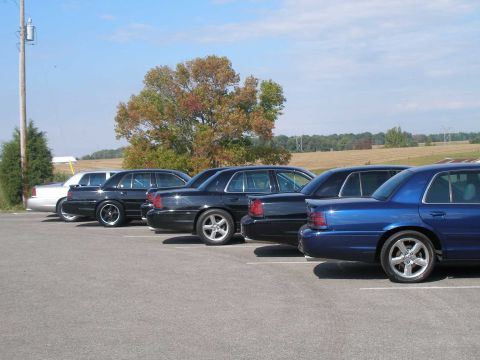 1998 Ford Crown Victoria - Rear three-quarter view along a line of full-size Ford Panther-platform sedans angle-parked in a rural lot, nearest a dark blue Mercury Mar…