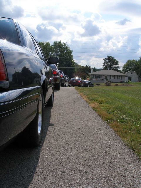 1998 Ford Crown Victoria - Low ground-level shot along the side of a black Ford Crown Victoria sedan parked on a gravel driveway, its polished flank and lace-style al…