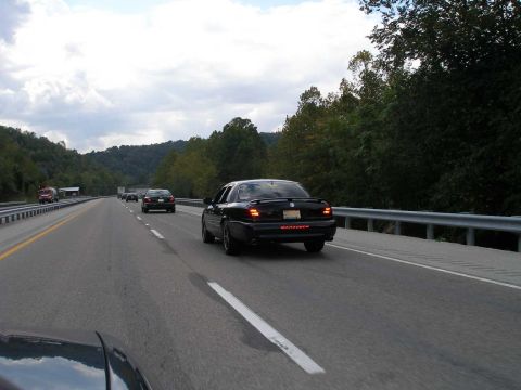 1998 Ford Crown Victoria - A black Mercury Marauder sedan driving away on a wooded highway, seen from behind with the 'MARAUDER' badge across the trunk and brake ligh…