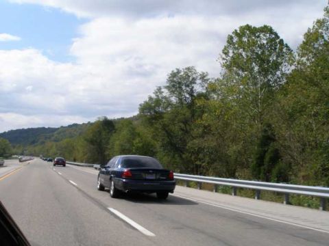 1998 Ford Crown Victoria - Rolling shot taken from another vehicle of a dark blue Ford Panther-platform sedan driving away down a divided highway lined with guardrail…