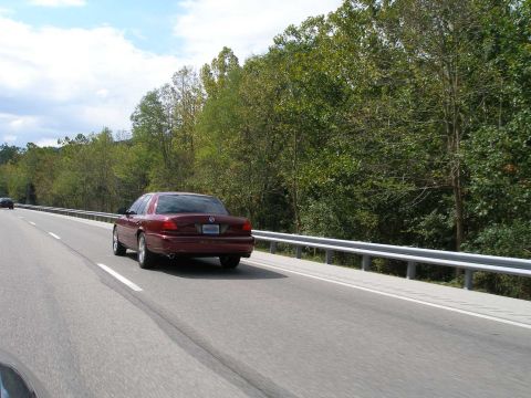 1998 Ford Crown Victoria - Rolling shot of a dark red Mercury Marauder full-size sedan photographed from behind on a divided highway, guardrail and green wooded hills…